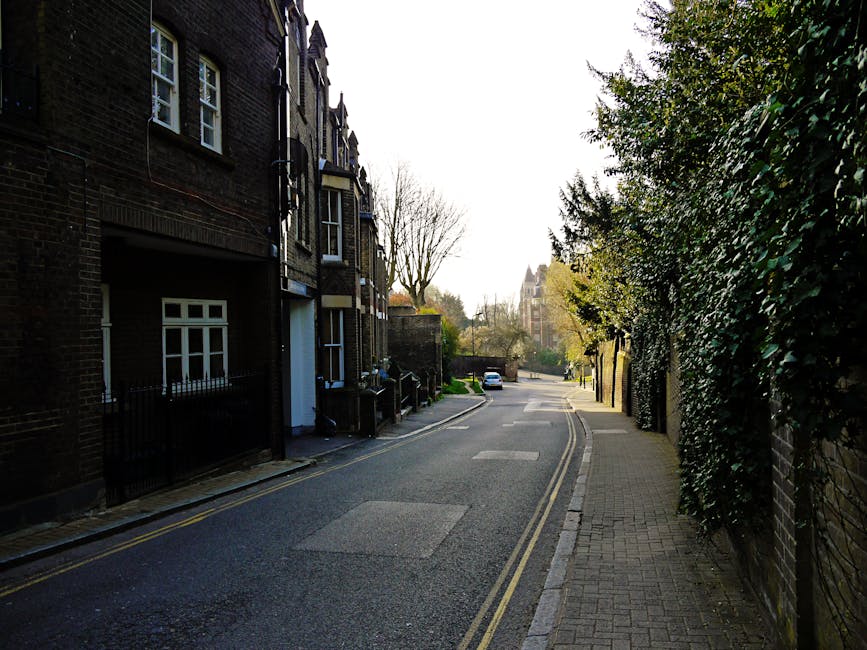A narrow urban alleyway flanked by brick buildings on both sides, leading towards a residential area with multiple back entrances, fire escapes, and garage doors. The pavement features double yellow l