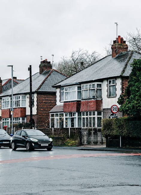 A row of Victorian-era terraced houses with ornate white balconies and large bay windows, situated on a sloped street in daylight under a partly cloudy blue sky. The houses have brick facades with dec